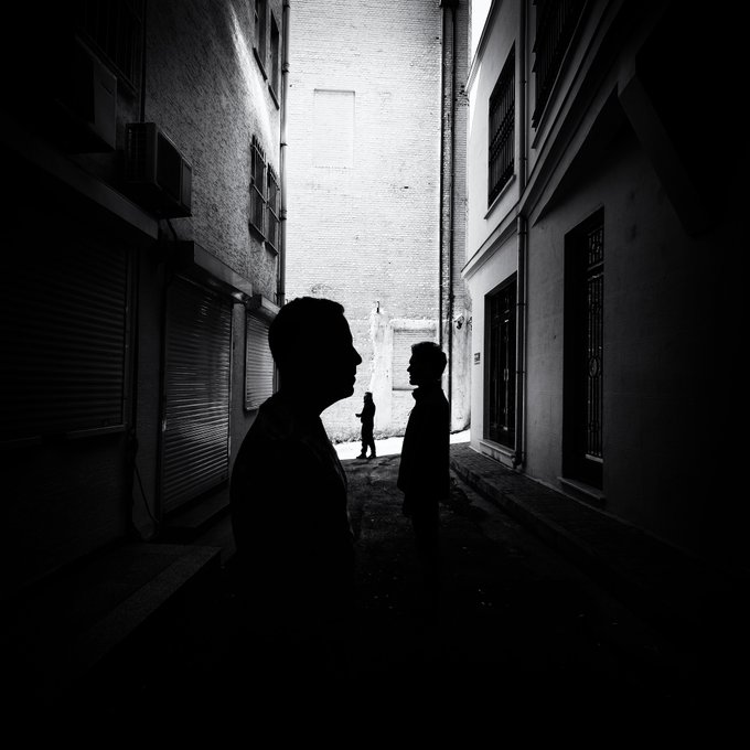 Silhouetted figures stand in a narrow, shadowy alleyway illuminated by a striking backlight, creating a dramatic interplay of dark foreground and bright background. The moody black-and-white composition, with its strong contrasts and anonymous forms, suits the playful mystery of "Guess who took this photo?"—perhaps the shadowy figure in the foreground is the unseen photographer themselves.