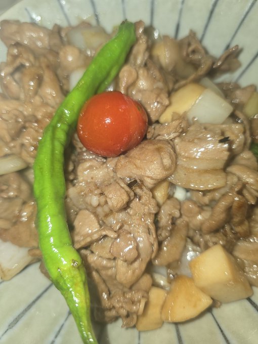 A close-up of a bowl of stir-fried meat and vegetables featuring a green chili pepper, a cherry tomato, onions, and potatoes over rice.