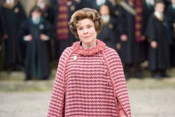 A woman with short curly hair wearing a textured pink poncho and a brooch, standing in what appears to be a Harry Potter film setting with figures in dark robes in the background.