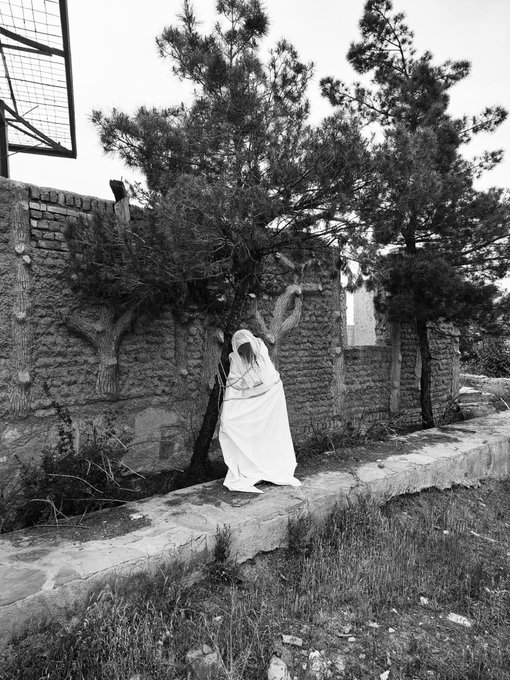 A black and white photograph of a person dressed in a long white robe and headscarf standing among trees next to a textured stone wall.
