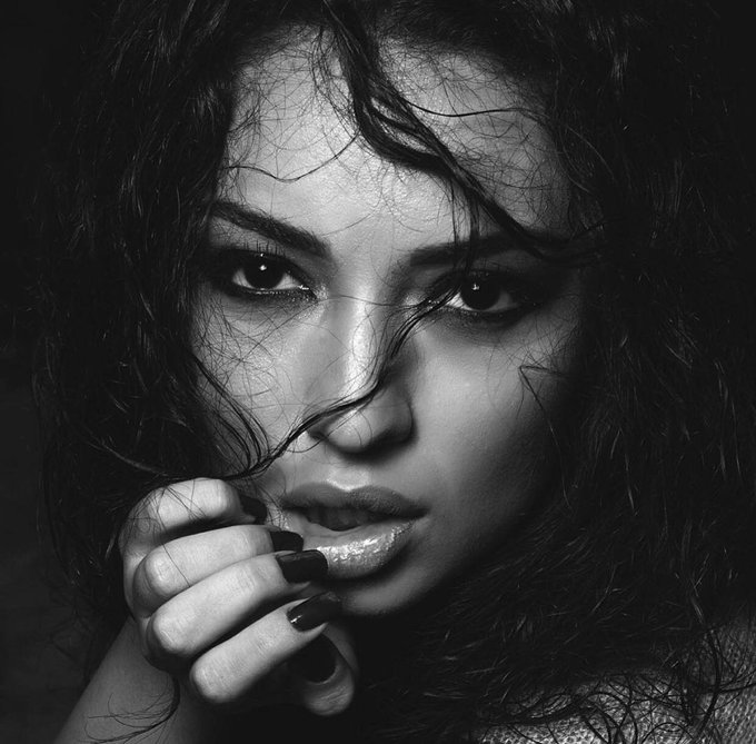 A high-contrast black and white close-up portrait of a woman with dark, messy hair, her hand resting near her face with dark painted nails.