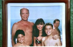 A framed vintage photograph showing a group of people, including a shirtless bald man and a woman in a bikini, posing together at a beach.