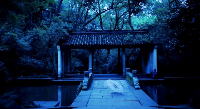 A serene, blue-toned nighttime photograph of a traditional Asian pavilion with a tiled roof, approached by a stone pathway crossing over water and surrounded by lush forest.