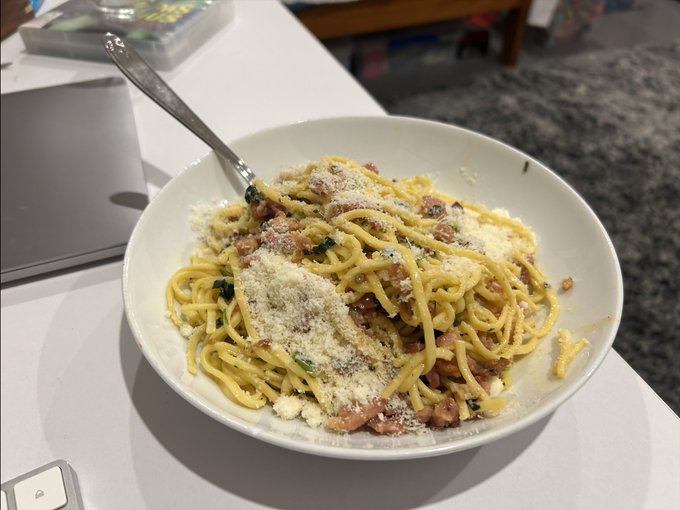 A bowl of spaghetti carbonara topped with grated Parmesan cheese and herbs, served on a white table with a fork.
