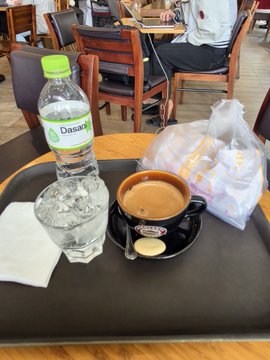 A tray on a cafe table holding a Dasani water bottle, a cup of coffee or tea with a cookie, and a plastic bag with what appears to be pastries.