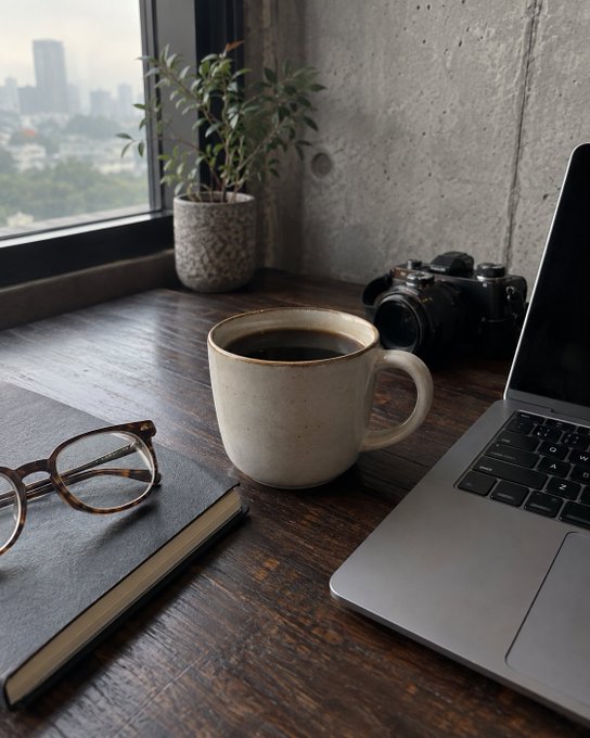 A still-life photo on a wooden desk featuring a cup of black coffee, an open laptop, a notebook with reading glasses, a vintage camera, and a potted plant by a window overlooking a city.