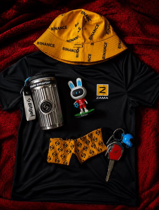A flat-lay arrangement of Binance-branded merchandise, including a yellow bucket hat, a black t-shirt with 'ZAMA' logo, a metal tumbler, a bow tie, and a keychain.
