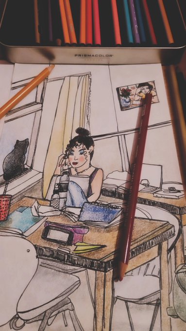 A hand-drawn sketch on paper illustrates a girl sitting at a desk with a laptop, accompanied by a black cat; colored pencils and a tin of Prismacolor pencils are visible nearby.