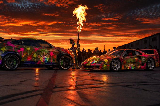 Two brightly graffiti-covered cars, including one labeled '34', face off at sunset on a runway, with a person holding a flaming torch between them.