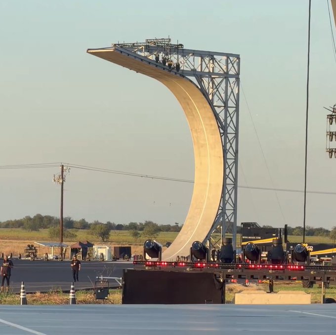 A massive half-pipe ramp structure with a curved wooden surface is being constructed outdoors on a flatbed trailer, supported by a tall metal scaffolding tower under a clear sky.