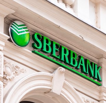 An illuminated green "SBERBANK" logo and emblem are mounted on the facade of an ornate building entrance, visually anchoring the news that Russia's banking giant is expanding crypto trading access to its massive 100+ million customer base.