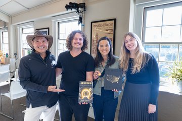 A group photo of four people smiling together indoors, with two of them holding up a book and a plaque.