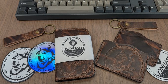 The image features a wooden desk with various items, including a keyboard, wallet, keychains, stickers, and a coin-shaped charm. The wallet has a lion charm on it, while the keychains have different designs. There are also several stickers scattered around the desk, some of which feature logos or text related to Dogecoin. A post on social media reads: "Dogecoin is money! Thank you for your support!". This message seems to be related to the wallet and keychains in the image, as they both have lion charm designs.