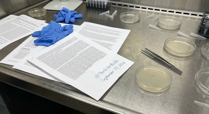 A laboratory bench setup with blue nitrile gloves, printed research documents, petri dishes, and tweezers, with one paper handwritten '@TheMicroBioHills September 27, 2026'.