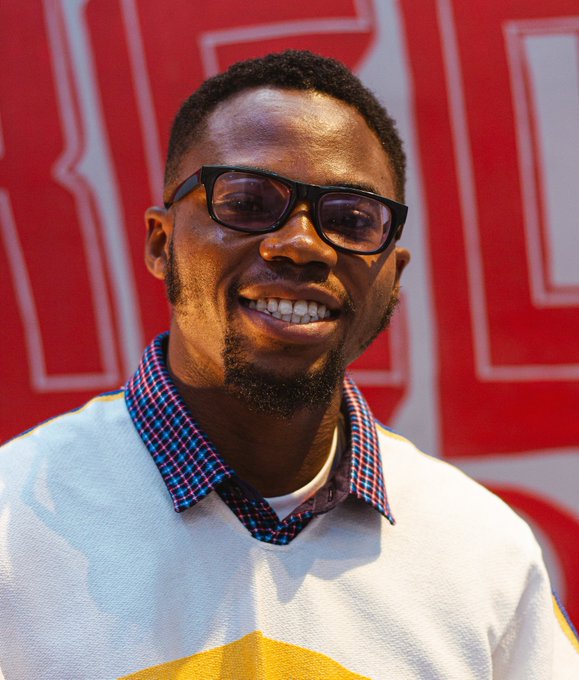 A smiling man wearing black glasses, a yellow sweater, and a blue-and-red checkered collar stands in front of a red backdrop with white letters.