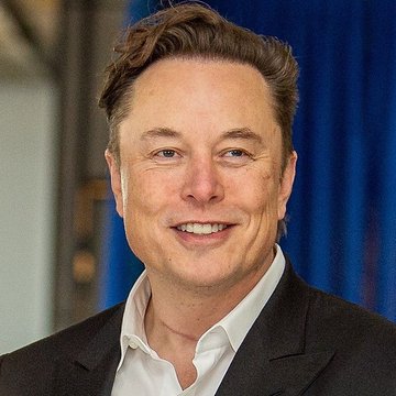 A portrait photo of Elon Musk smiling, wearing a black suit jacket and white shirt against a blue backdrop.