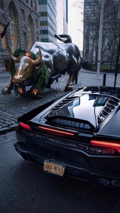 A black sports car with a New York license plate reading 'ORO NERO' parked on a cobblestone street in front of the iconic bronze Wall Street Charging Bull statue, which is decorated with a green Christmas wreath.