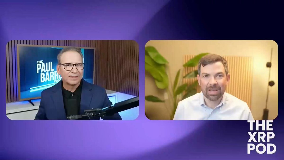 The image shows two men sitting across from each other at a table, engaged in an interview or discussion. They are both wearing glasses, suggesting that they might be experts in their field. The background of the image features a purple wall with a television screen mounted on it, which is displaying the names "Paul Barlow" and "The XPOD". This suggests that the men could be discussing topics related to blockchain technology or finance.