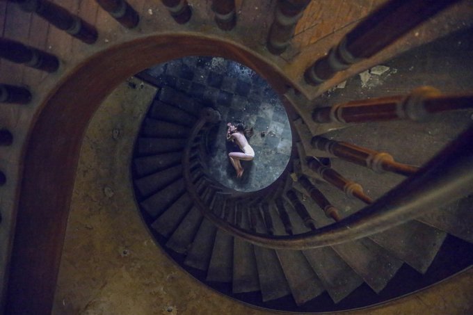 An overhead photograph of a person curled up at the bottom of a spiral staircase with ornate stone balusters.