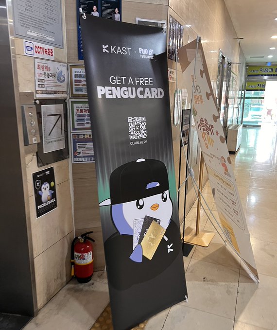 A black promotional banner for 'KAST x Pudgy Penguins' advertising 'GET A FREE PENGU CARD' with a QR code and a penguin character wearing a cap, displayed in a building lobby near a CCTV sign and fire extinguisher.