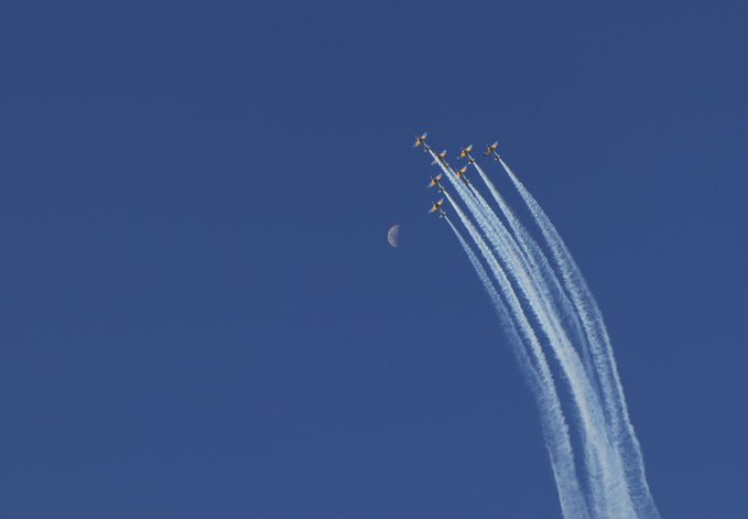 A formation of six jets flies through a deep blue sky leaving white contrails, with a crescent moon visible to the left.