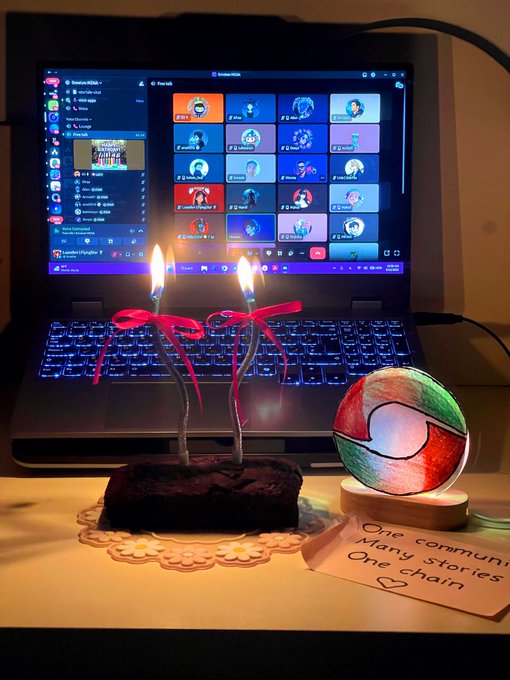 A laptop displays a Discord server, with a birthday celebration in front featuring two lit candles, a small chocolate cake, and a handwritten note reading 'One community, Many Stories, One chain'.