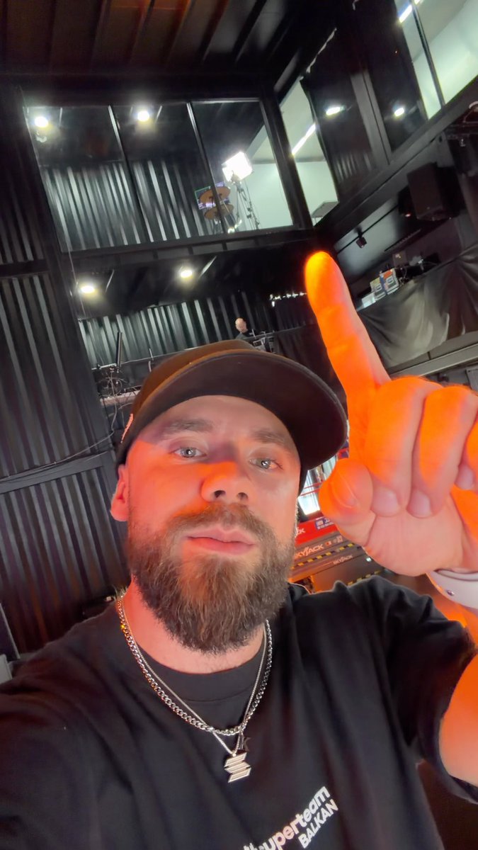 A bearded man wearing a black cap and a 'Hyperteam BALIKAN' t-shirt points upward in an indoor setting with dark, industrial-style ceiling fixtures.