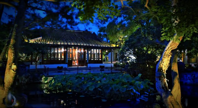 A nighttime photograph of a traditional East Asian building with glowing windows, reflected in a pond surrounded by trees and lotus plants.