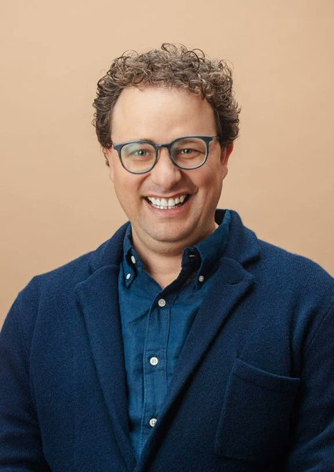 urn of water, a man with curly hair and glasses, wearing a blue jacket against a beige background.