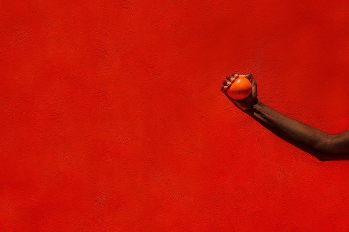 A hand holds a small orange object against a vibrant red textured wall, with the arm extending from the right side of the frame.