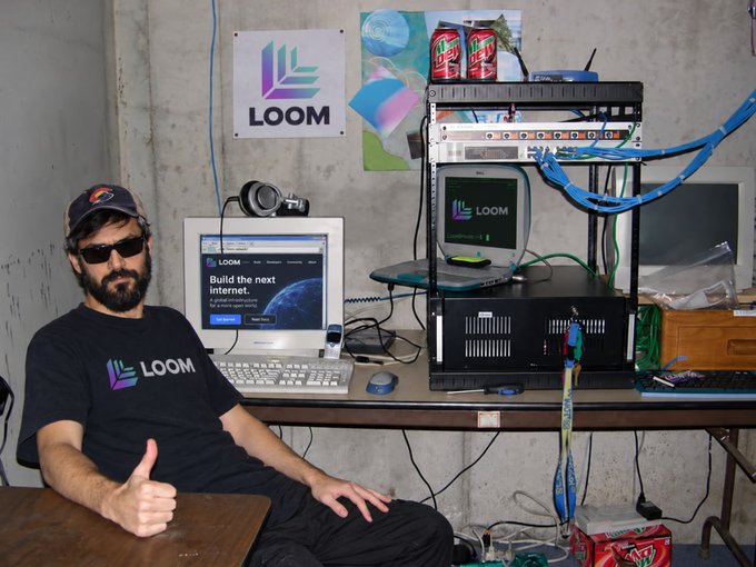A bearded man wearing sunglasses and a "LOOM" branded t-shirt giving a thumbs up, seated next to a workstation with computer equipment displaying the LOOM logo.