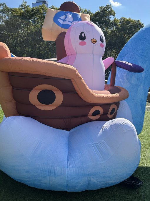An outdoor inflatable display features a pink penguin character sitting in a brown pirate ship with a white sail bearing a blue logo.