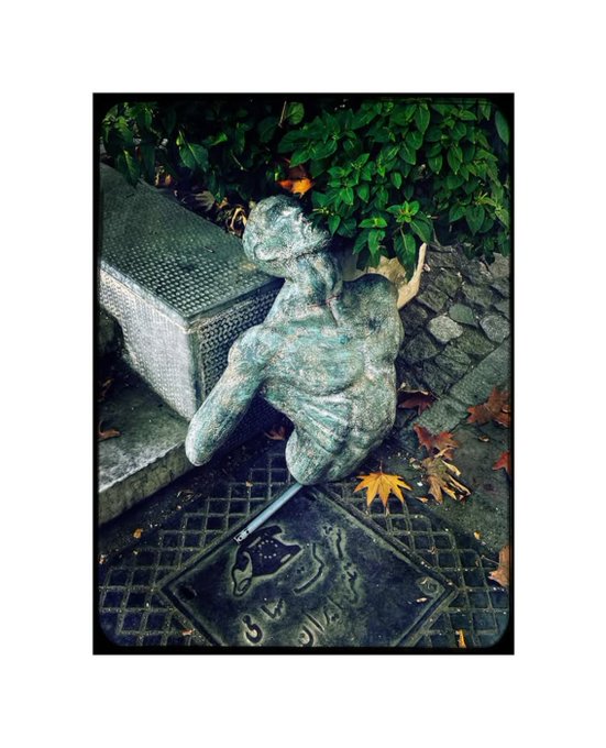 A photograph of a weathered bronze statue of a man with his arm raised, positioned next to a stone pedestal with autumn leaves scattered around.