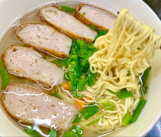 A bowl of Asian noodle soup with clear broth, yellow noodles, green vegetables, and several slices of meat loaf or fish cake.