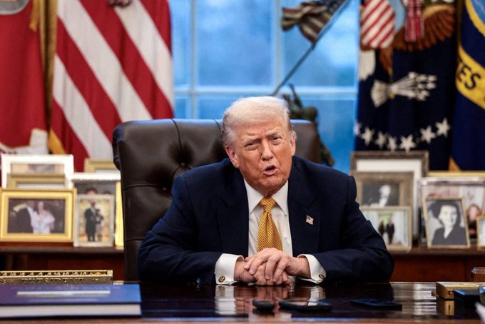 Donald Trump sits at a desk in the Oval Office, wearing a yellow tie, with the American flag and a flag displaying 'ES' visible behind him.