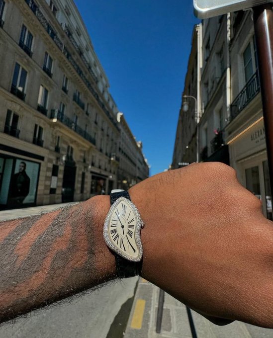 A first-person view of a wrist wearing a triangular diamond-encrusted watch on a black strap, set against a Parisian street with classic Haussmannian architecture.