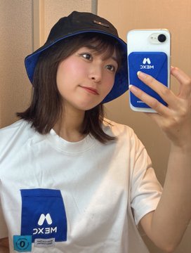 A young woman takes a mirror selfie while wearing a navy blue "MEXC" bucket hat and matching white t-shirt featuring the MEXC exchange logo, with her phone case also displaying the "MEXC" branding—visually reinforcing the post's reference to MEXC as a remaining crypto trading option in Japan after Bitget's restriction.