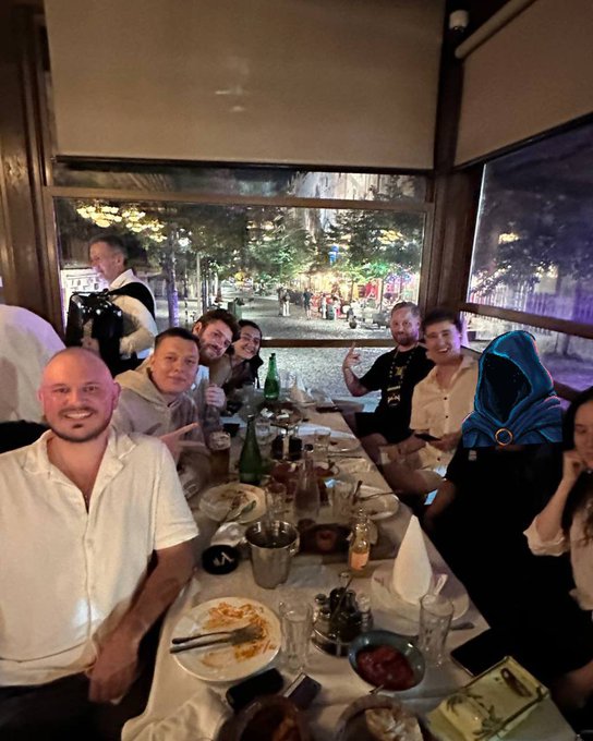 A group of people smile and pose for a photo while gathered around a long dining table in a restaurant at night.