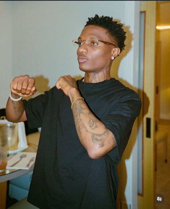 A young man with short dreadlocks, glasses, and tattoos wearing a black t-shirt, posing with his fists raised indoors.
