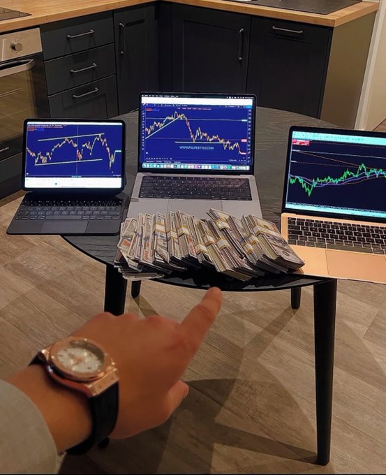 A photo of three laptops on a small round table displaying trading charts, with stacks of cash in front and a person's hand pointing with a luxury watch visible.