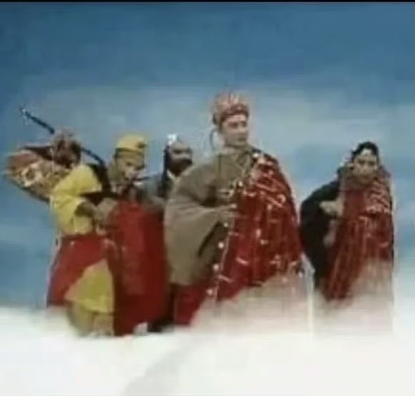 A vintage scene, likely from a Chinese opera or fantasy TV show, showing several characters in elaborate traditional costumes including a monkey king figure with a staff, standing on clouds against a blue sky.
