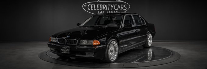A black BMW sedan is displayed on a rotating platform at Celebrity Cars Las Vegas, as indicated by the logo at the top of the image. The car is the one Tupac was fatally shot in, which is now listed for auction alongside the renewed public interest in his case.
