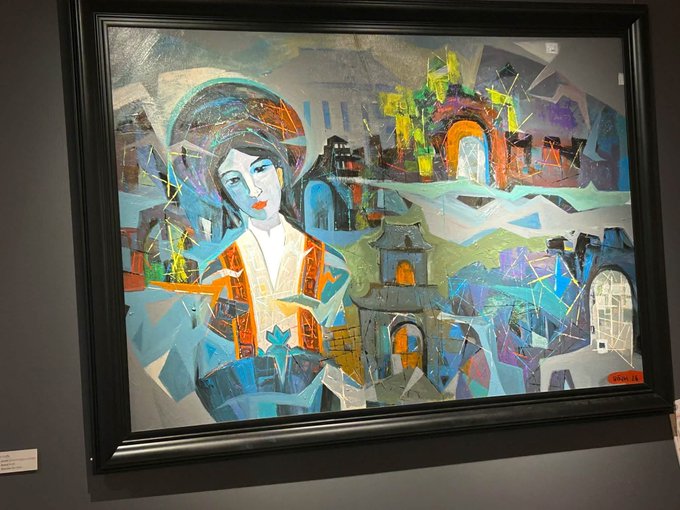 A framed abstract painting depicting a woman in traditional dress against a colorful, cubist-style background of buildings and landscapes, displayed on a dark wall.