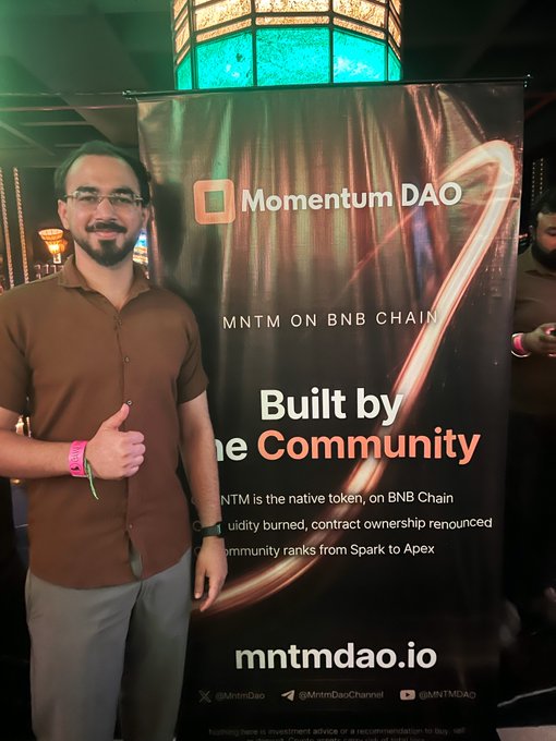 A man with a beard and glasses gives a thumbs-up next to a banner for "Momentum DAO" ("MNTM ON BNB CHAIN", "Built by the Community", "mntmdao.io").