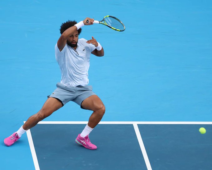 A tennis player in a white shirt, gray shorts, and bright pink shoes lunges in a wide stance to return a yellow tennis ball with his racket on a blue hard court.