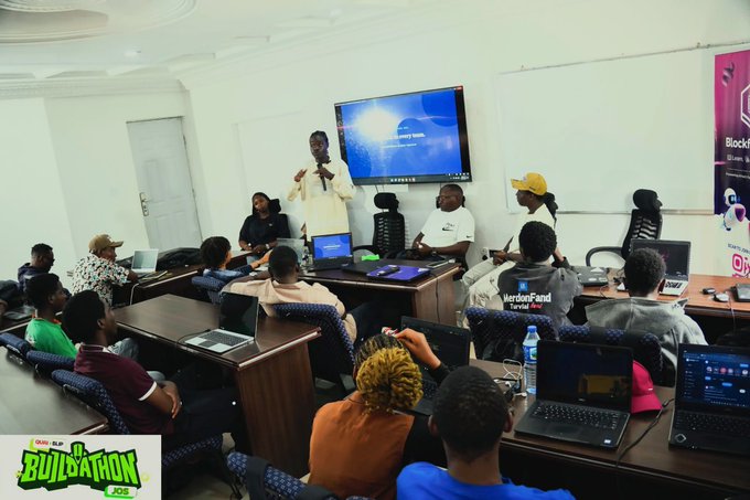 A presenter addresses a group of attendees seated at desks with laptops during a workshop, with a 'Jos BLOCKATHON' logo visible in the bottom left corner.