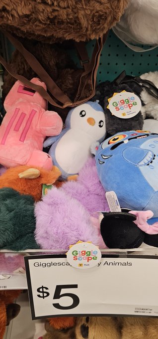 A store shelf display of 'Gigglescape' plush animals priced at $5, featuring various colorful stuffed toys including a pink elephant, penguin, blue monster, and a black character.