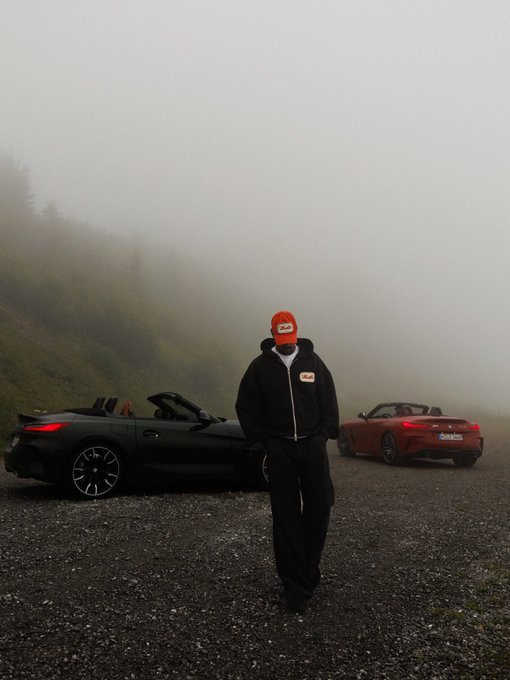 A person in a black tracksuit and red cap stands in foggy conditions between a black and a red convertible sports car parked on a gravel road.