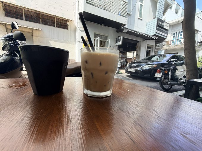 An iced coffee in a glass and a black ceramic cup sit on a wooden table at an outdoor café, with a parked car and motorcycle visible across the street.