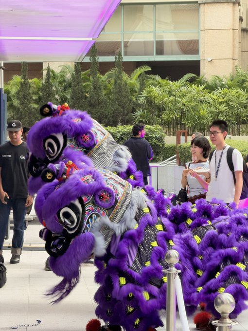 The image shows a vibrant purple and yellow Chinese lion dance costume being performed outdoors, with spectators including event security watching the traditional cultural performance.